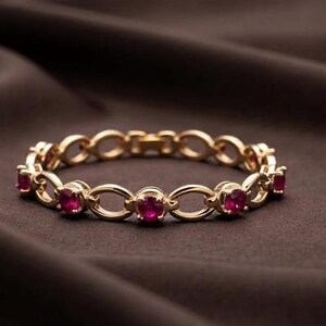 May include: A gold-toned metal bracelet featuring a series of interconnected oval links alternating with small, round, vibrant red gemstones set into the metal frame, creating a continuous circular design with a secure clasp mechanism.