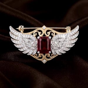 May include: A brooch featuring a central rectangular deep red gemstone set in a gold-toned filigree frame. Two silver-toned wings encrusted with small clear stones extend from the center. A horizontal pin mechanism is visible at the top.