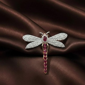 May include: A dragonfly brooch with a gold body, ruby accents, and diamond-studded wings. The brooch is set against a dark brown, satin-like background. The dragonfly's body is adorned with a series of rubies, and the wings are covered in small, sparkling diamonds.