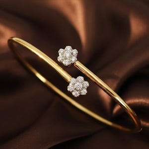 May include: A gold-colored bangle bracelet with two flower-shaped accents. Each flower is adorned with small, sparkling gemstones. The bracelet curves gracefully, creating an elegant and delicate design. The background is a rich, brown fabric.