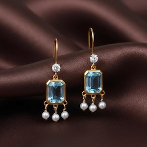 May include: A pair of gold-colored drop earrings. Each earring features a rectangular, light blue gemstone, a small, round, clear gemstone, and three small, white pearls. The earrings are displayed against a brown background.