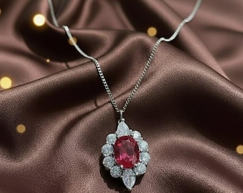 May include: A silver necklace with a pendant featuring a large oval ruby surrounded by smaller, clear gemstones. The ruby is a deep red color, and the necklace chain is delicate. The pendant is set against a brown satin background.