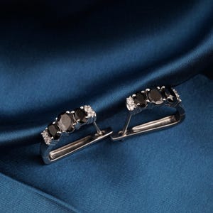 Puede incluir: Un par de pendientes plateados con diseño rectangular. Cada pendiente presenta cinco gemas negras ovaladas y pequeñas piedras blancas, sobre un fondo de tela azul oscuro.