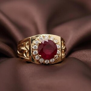 Può includere: Un anello d'oro con una grande gemma rosso scuro rotonda al centro, circondata da un alone di pietre chiare più piccole. L'anello presenta motivi floreali ornati sui lati.