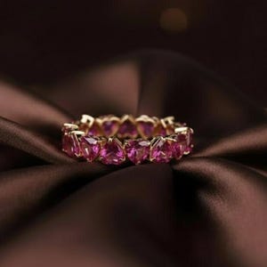 May include: A gold ring with a continuous row of heart-shaped pink gemstones. The ring is displayed on a dark brown, satin-like fabric, which enhances the sparkle of the gemstones and the gold setting.