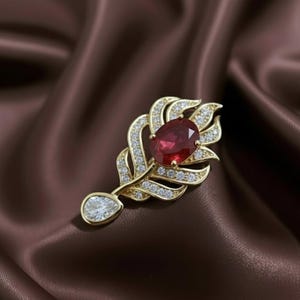 May include: A gold-toned brooch in a feather design, featuring a large oval ruby gemstone at the center. The feather is adorned with small, sparkling clear gemstones. A pear-shaped clear gemstone hangs at the bottom of the feather.