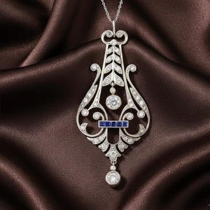 Puede incluir: Un collar con colgante de plata ornamentado con un diseño de filigrana detallado. El colgante presenta piedras preciosas claras y un acento rectangular de piedra preciosa azul. Una piedra preciosa redonda cuelga en la parte inferior. El collar se muestra sobre una tela de satén marrón.