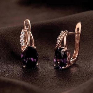 May include: A pair of rose gold hoop earrings featuring oval-cut deep purple gemstones at the base and a curved accent bar set with small clear stones along the front. The earrings have a hinged closure mechanism and a polished metallic finish.