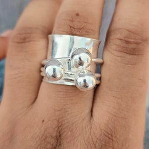 May include: A silver ring with a wide band and three small, spherical accents. The ring is worn on a finger, showcasing its polished, reflective surface. The design is simple yet eye-catching, suitable for everyday wear.