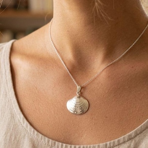 Könnte beinhalten: Eine silberne Muschel-Anhänger-Halskette. Der Anhänger ist ein detailliertes, dreidimensionales Muscheldesign. Die Halskette wird von einer Person getragen, die ein beiges Tanktop trägt. Der Hintergrund ist unscharf.