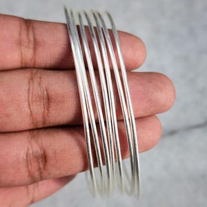 Könnte beinhalten: Ein Stapel dünner, silberfarbener Armreifen. Die Armreifen sind rund und scheinen aus Metall zu sein. Das Bild ist eine Nahaufnahme, wobei die Armreifen in einer Hand vor einem neutralen Hintergrund gehalten werden. Die Armreifen sind aufeinander gestapelt.