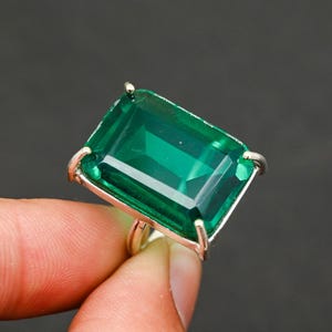 May include: A silver ring featuring a large, rectangular emerald-green gemstone. The gemstone is set in a silver band with four prongs holding it in place. The ring is displayed against a dark background.