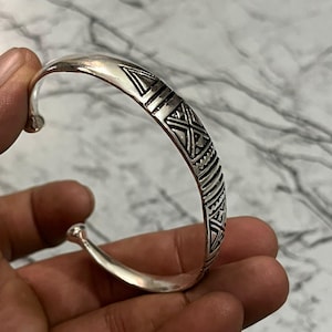 May include: A silver cuff bracelet with an open design. The bracelet features geometric patterns, including triangles and lines, etched along the top. The ends of the bracelet are rounded. The bracelet is held in a hand.