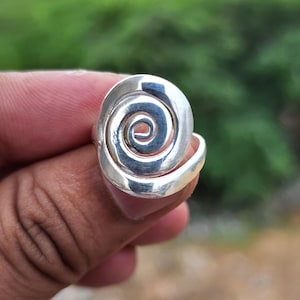 May include: A silver-colored ring with a spiral design. The ring features a raised, swirling pattern that creates a three-dimensional effect. The ring is held up against a blurred green background.