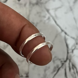 Op de afbeelding: Een zilveren ring met een dubbel bandontwerp. De ring wordt om een vinger gedragen en toont zijn gladde, reflecterende oppervlak. De banden liggen dicht bij elkaar en creëren een unieke, minimalistische stijl. De ring lijkt van glanzend metaal te zijn gemaakt.