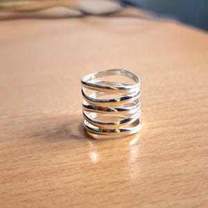Könnte beinhalten: Ein silberner Ring mit einem gestapelten Design. Der Ring hat fünf Bänder aus poliertem Metall, wodurch ein geschichteter Effekt entsteht. Die Bänder sind gleichmäßig verteilt und haben eine glatte, reflektierende Oberfläche. Der Ring ist ein schlichtes, aber elegantes Schmuckstück.
