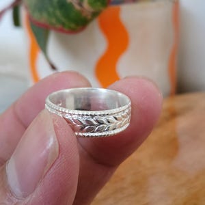 May include: A silver ring with a detailed leaf pattern. The ring is held between fingers, showcasing its intricate design. The ring's band features a repeating leaf motif, adding a touch of nature-inspired elegance to the jewelry.