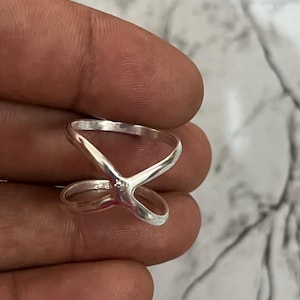 Peut inclure: Une bague en argent avec un design en forme de X. La bague a un petit ornement rond au centre du X. La bague est tenue dans une main.