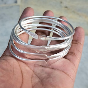 Pode incluir: Um conjunto de pulseiras de prata com textura martelada. As pulseiras abertas são lisas e brilhantes, refletindo a luz. As pulseiras são seguradas em uma mão, mostrando seu design e artesanato.