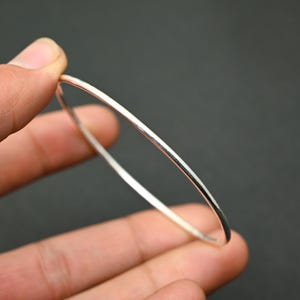 May include: A slender, silver-coloured bangle bracelet. The minimalist, round design is held by a hand against a dark background. The bracelet appears to be made of a shiny metal.
