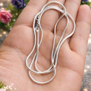 Op de afbeelding: Een zilveren slangketting met een karabijnsluiting wordt getoond in een open handpalm. De ketting is gemaakt van kleine, onderling verbonden schakels die een gladde, flexibele textuur creëren. De achtergrond toont onscherpe bloemen.