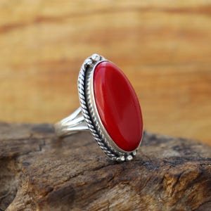 Op de afbeelding: Een zilveren ring met een grote, ovale, helderrode steen. De steen is gezet in een gedetailleerde bezel met een gedraaid touwontwerp rond de rand. De ring wordt gepresenteerd op een houten oppervlak.