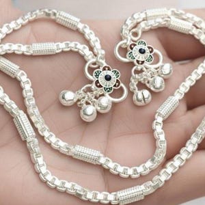 May include: Silver anklet with a chain-link design. The anklet features decorative flower charms with red, green, and black accents, along with small dangling bells. The jewelry is displayed on a light background.