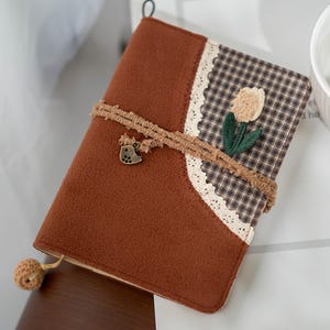 Puede incluir: Un diario marrón con un diseño floral decorativo y un cierre tejido. El diario presenta un patrón de cuadros, ribete de encaje y un pequeño dije de pájaro. Una pequeña borla redonda cuelga de la parte inferior.