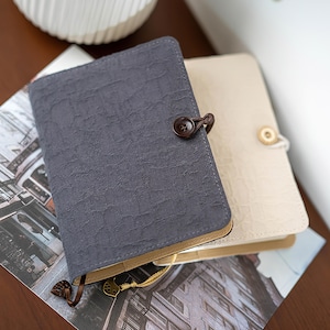 May include: Two textured notebooks, one gray and one cream, are stacked on a surface. The gray notebook has a brown button closure. The cream notebook has a light-colored button closure. Both notebooks have a ribbon bookmark. The notebooks are on top of a printed image.