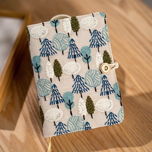 May include: A beige fabric journal cover with an embroidered forest pattern in shades of blue, green, and white. The cover features a wooden button closure and a gold-colored bookmark ribbon. The design includes various tree styles.
