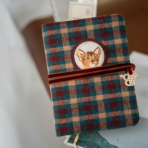 May include: A plaid journal with a cat embroidery patch. The journal features a teal, brown, and burgundy plaid pattern. A burgundy velvet ribbon with a pearl and rhinestone crown closure secures the journal.