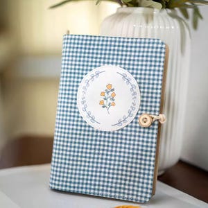 Puede incluir: Un cuaderno con un patrón de cuadros vichy azul y blanco. La portada tiene un parche redondo blanco con un diseño floral y un borde de hojas. Un botón de madera y una cuerda se utilizan para cerrar el cuaderno. El cuaderno mide unos 15 cm de alto.
