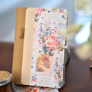 May include: A vertical notebook featuring a brown kraft paper cover and a cream-colored fabric panel decorated with a floral print of pink roses, blue flowers, and a cottage scene. The cover includes cursive text and a wooden button closure.