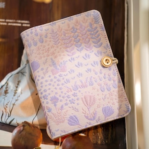 May include: A floral patterned journal with a button closure. The cover features a repeating design of lavender and pink flowers and foliage on a cream background. The button is made of wood and is attached with a beige cord.