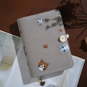 May include: A rectangular gray fabric notebook cover featuring embroidered cat faces in shades of brown, gray, and white. The cover includes small brown paw print accents, a white button closure with a cord loop, and a small metal cat-shaped charm.
