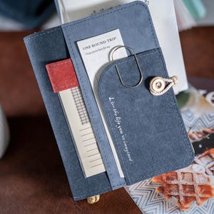 May include: A blue suede-effect journal with a flap closure and a decorative button. The journal contains a paper clip, a small notepad, and a bookmark. The text "ONE ROUND TRIP I hope you're here for me" is visible on a white card. The text "Live the life you've imagined" is printed on the flap.