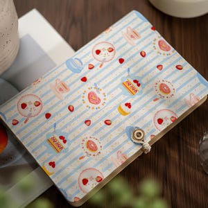 May include: A rectangular notebook featuring a light blue and white striped cover with a repeating pattern of strawberries, teacups, donuts, and cake slices. The cover is secured with a small, round wooden button and a white cord loop closure.