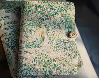 May include: A floral-patterned journal with a button closure. The cover features a cream-colored background with a pattern of pink roses, green foliage, and blue accents. The journal is closed with a wooden button and a loop.