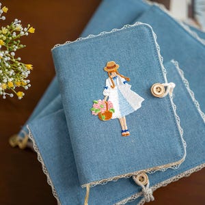 May include: Blue denim journal with embroidered illustration of a girl in a dress and hat carrying a basket of flowers. The journal has a lace trim and a wooden button closure. Several journals are stacked.