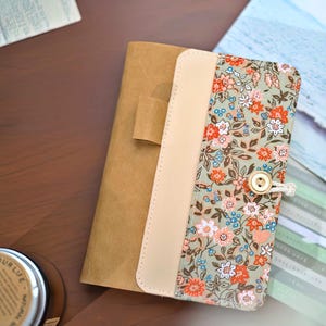 Może przedstawiać: Okładka na dziennik z kwiatowym nadrukiem i zapięciem na guzik. Okładka posiada grzbiet w kolorze beżowym, przypominającym skórę oraz kwiatowy wzór w odcieniach pomarańczu, różu i błękitu na zielonym tle. Dziennik jest częściowo otwarty.