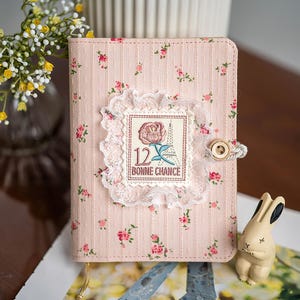 May include: A pink journal with a floral pattern and a decorative lace-trimmed patch featuring a rose and the text "12 Bonne Chance." The journal has a button closure and a small bunny figurine is next to it.