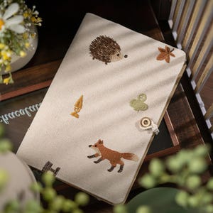 May include: A cream-colored fabric book cover featuring embroidered motifs of a brown hedgehog, a rust-colored fox, a brown maple leaf, a green clover, and a yellow acorn. The cover includes a circular wooden button closure with a white cord loop.