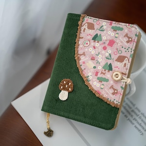 May include: A green corduroy journal with a pink floral cover featuring houses, trees, and sewing tools. A mushroom applique and a bird charm dangle from the journal. The journal is closed with a button and loop.