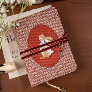 Puede incluir: Un diario decorativo con una cubierta tejida roja y blanca. El diario presenta un diseño de conejo bordado en un óvalo rojo, una cinta de terciopelo burdeos y un dije dorado. El diario está cerrado y está sobre una superficie de madera.