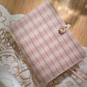 Puede incluir: Un diario cerrado con una cubierta a cuadros rosa y blanca, asegurado con un botón de madera y un cierre de cuerda. El diario descansa sobre un tapete de encaje blanco, con marcadores de cinta color cobre.
