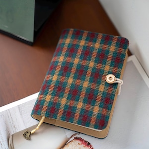 Puede incluir: Un diario con un patrón de cuadros con cierre de botón. La cubierta presenta un diseño en verde azulado, rojo y amarillo. Se ve un marcapáginas dorado. El diario está sobre una revista. Un accesorio elegante para escribir y tomar notas.
