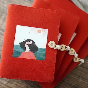 May include: A stack of three red fabric journals. Each journal has a small, square illustration of a woman with black hair and a red shirt. The journals are bound with a white string and wooden buttons.
