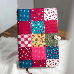 Puede incluir: Un diario de patchwork con una variedad de cuadrados de tela coloridos. La cubierta presenta cuadrados de telas rojas, azules, rosas y amarillas con patrones como lunares, cerezas y diseños florales. Un cierre de botón y cuerda asegura el diario.