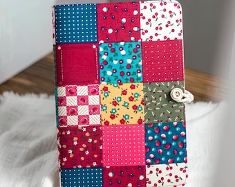 May include: A patchwork journal with a variety of colorful fabric squares. The cover features squares of red, blue, pink, and yellow fabrics with patterns like polka dots, cherries, and floral designs. A button and string closure secures the journal.