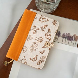 May include: A beige book cover with a butterfly and floral pattern, featuring an orange spine. The cover has a wooden button closure with a rope tie and a decorative charm. The book cover is on a wooden surface.